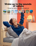 A person waking up and stretching next to a sunrise alarm clock with wake up light and various sound options.